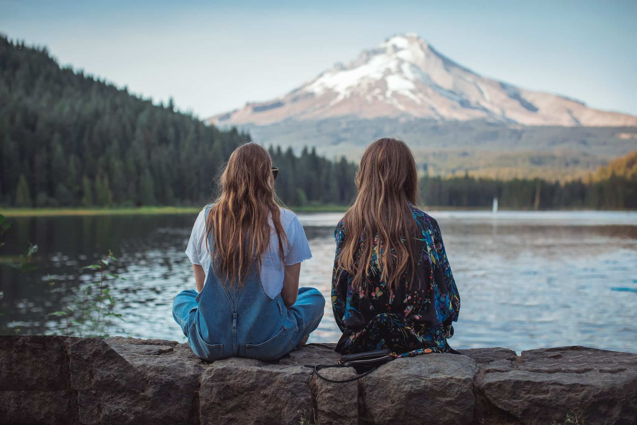 Two Women Sitting on Rock Facing on Body of Water and Mountain in oregon