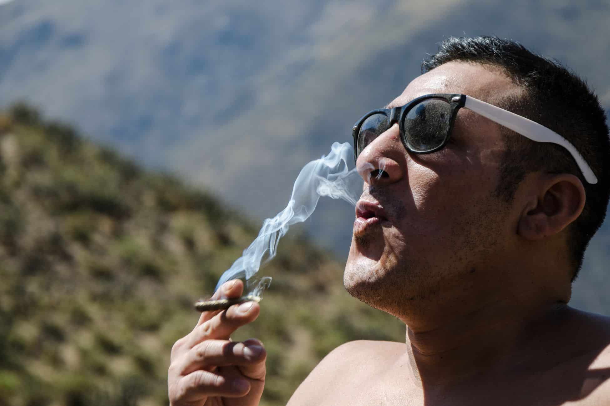 A man in sunglasses smoking a cigarette in the mountains, wondering if THCA is legal in Illinois.