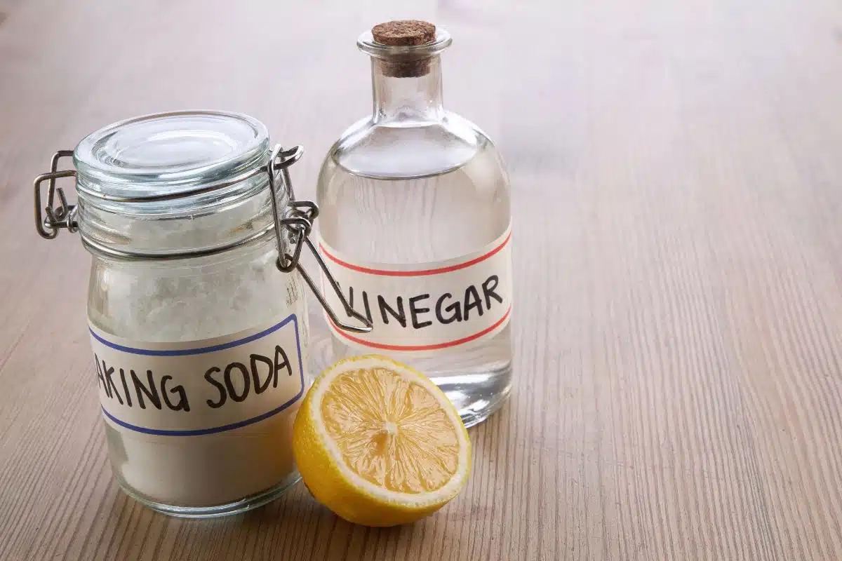 A jar of baking soda, a bottle of vinegar, and a half lemon on a wooden surface are natural cleaning solutions for vape juice on cloth
