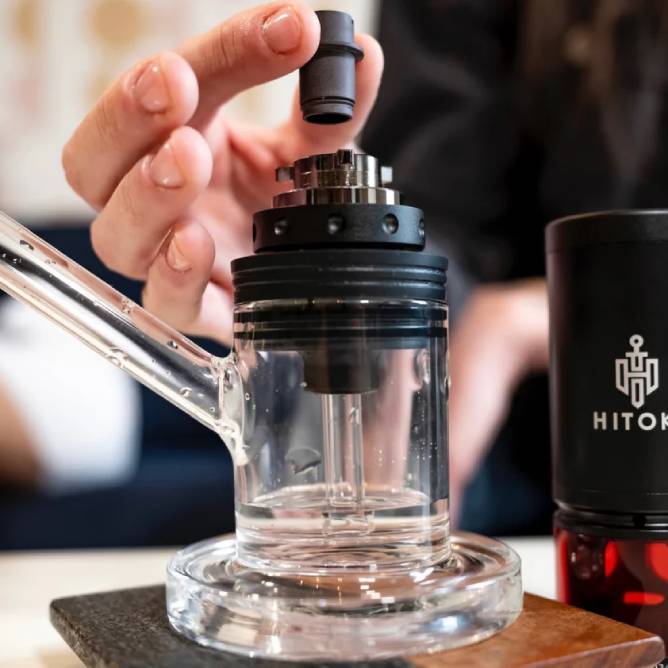 A person assembles the Hitoki Saber Laser Bubbler Kit, featuring a cylindrical glass body with a detachable black top. A hand attaches a small, cylindrical component to the device.