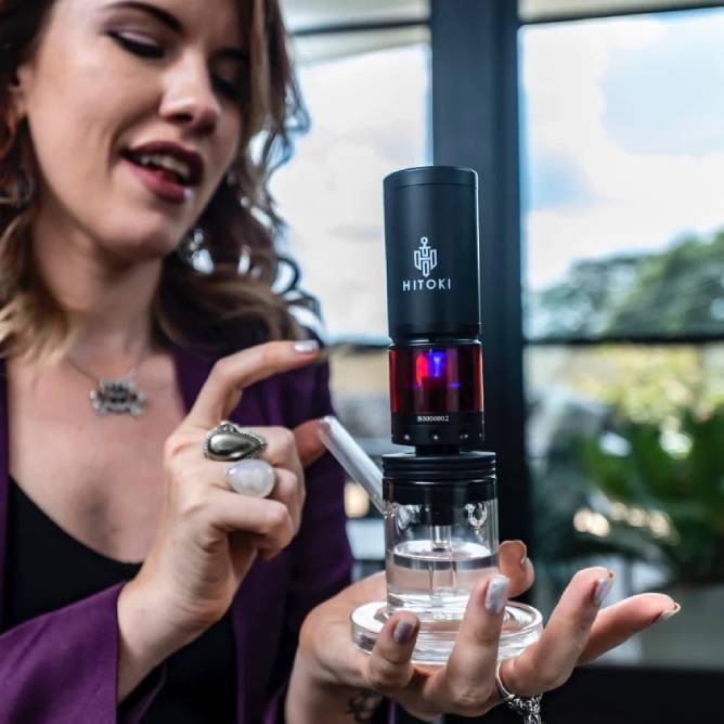 A woman holds a device labeled HITOKI, identified as the Hitoki Saber Laser Bubbler Kit, featuring a sleek black top and clear base. Smiling, she showcases its stylish design with plants and windows in the background.