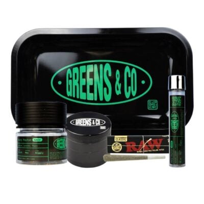 A collection of cannabis-related items including a branded black tray labeled Greens & Co, a container with green labeling, a grinder, a rolled joint in a glass tube, and RAW rolling papers.