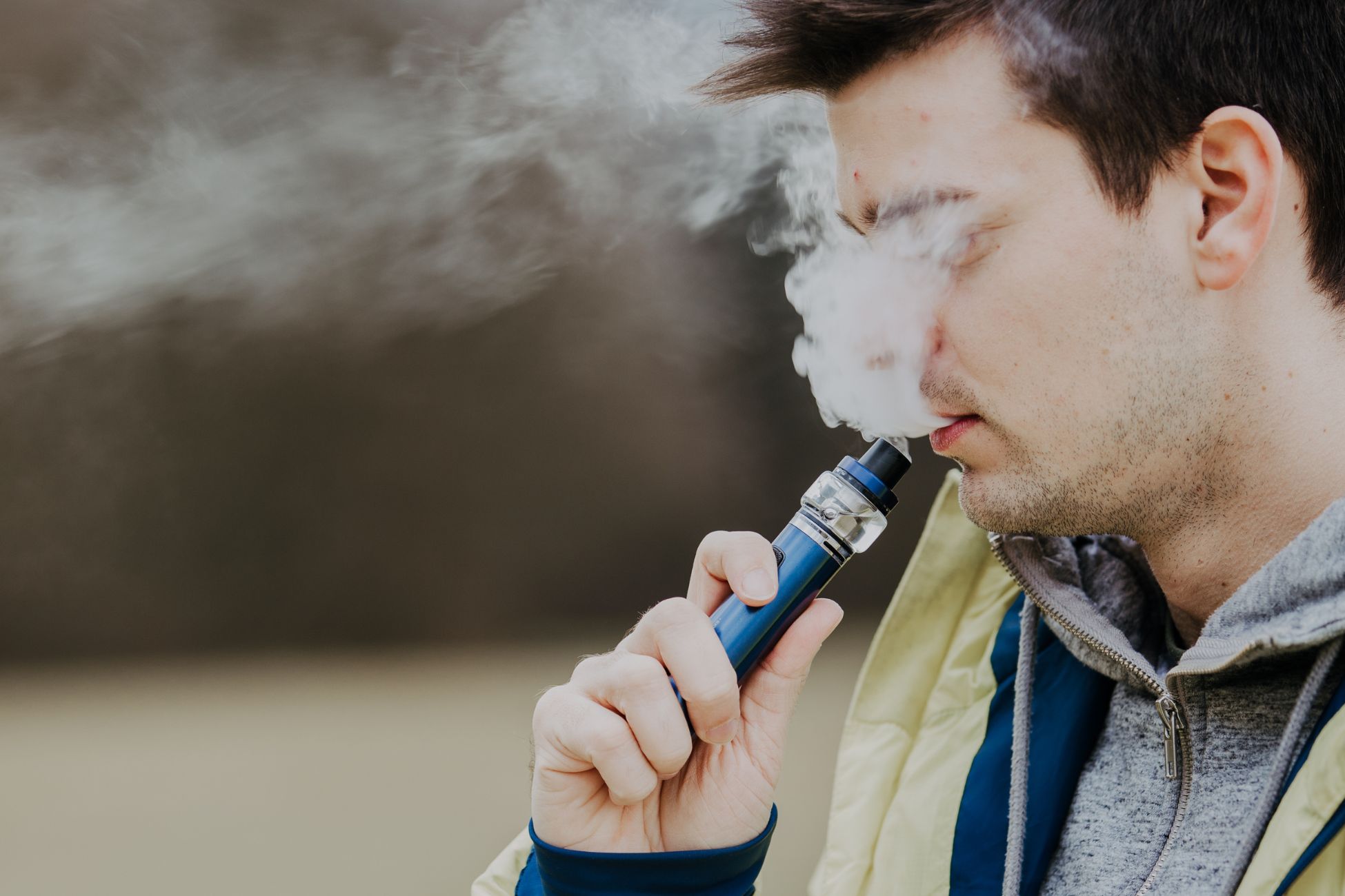 A person holding a blue vape pen, pondering how long a cart stays in your system, exhales a cloud of vapor. They wear a light jacket and hoodie, standing outdoors with a blurred background.