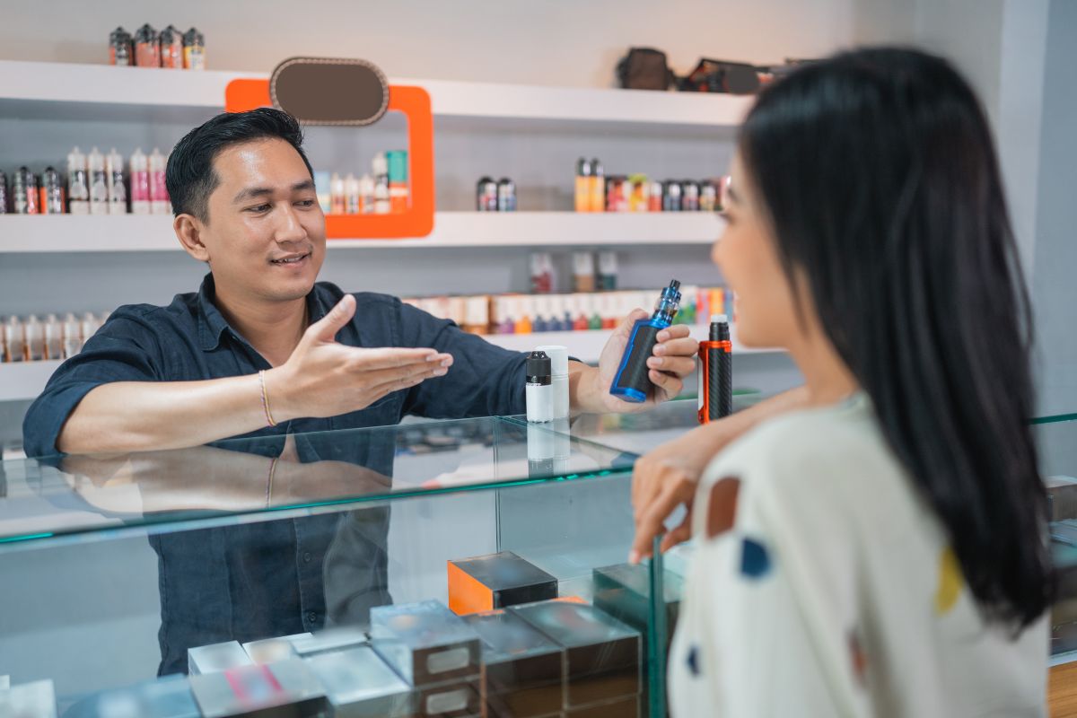 A man behind a glass counter in a vape shop discusses with a woman. Holding a vape device, he gestures animatedly. Shelves stocked with various Circle K Vapes products are visible in the background, enticing customers to consider making a buy.