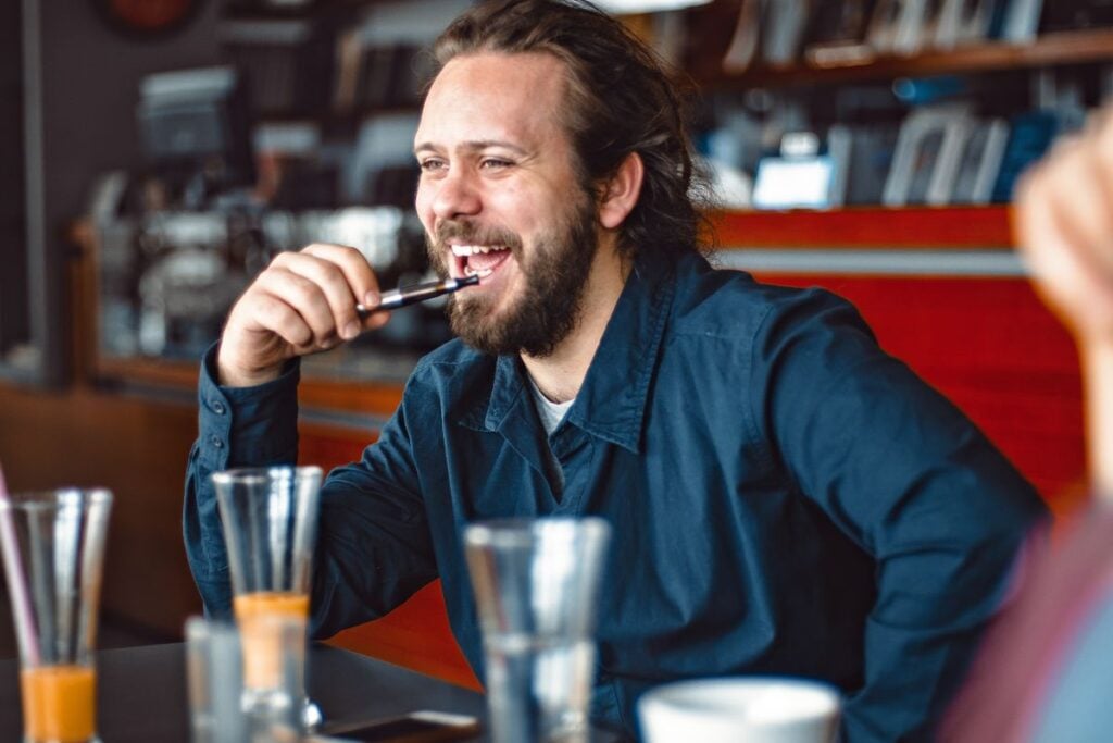 A man with a beard and long hair smiles while sitting at a table in a cafe. Holding a pen near his mouth, he wears a dark blue shirt. Beside him are glasses, an empty cup, and his sleek vape from Circle K Vapes resting on the table against the blurred backdrop of cozy bookshelves.